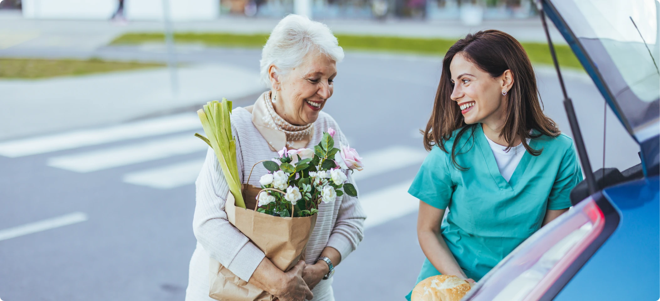 An older woman holding groceries and flowers, smiling with a woman beside a car.