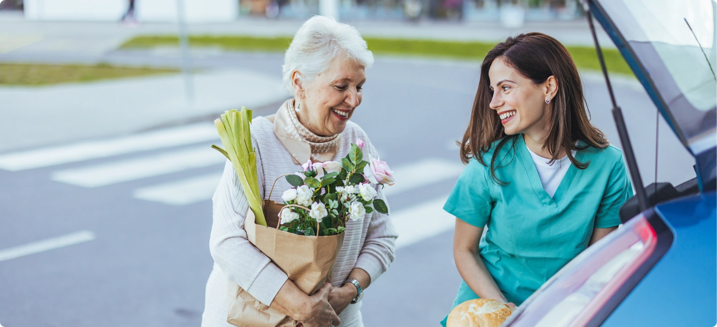 An older woman holding groceries and flowers, smiling with a woman beside a car.